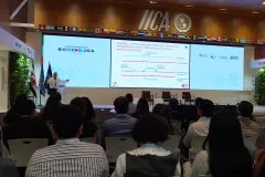 Un expositor habla desde un podio en el auditorio del IICA. En la pantalla se proyecta una diapositiva sobre cisgénesis; en la pared superior se ven las banderas de los países miembros del IICA.