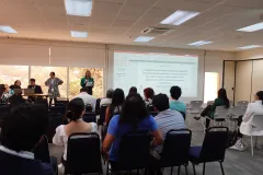 Sala de reuniones con asistentes sentados de espaldas, viendo una presentación proyectada sobre bacterias ácido lácticas agroindustriales y los hongos Aspergillus flavus y Fusarium verticillioides; al frente, una mujer habla con micrófono.